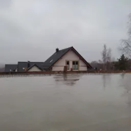 Świeżo wylana, mokra wylewka anhydrytowa na dachu z widocznymi elementami zbrojenia i odbiciem drzew na powierzchni; w tle domy jednorodzinne pod pochmurnym niebem.