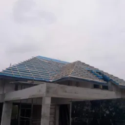 Surowy stan domu murowanego z widoczną konstrukcją dachu pokrytą niebieską folią, drewniane łaty przygotowane do dalszych prac dekarskich, pochmurne niebo w tle.