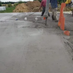 Wylewanie betonu na przygotowany teren pod budowę, widoczni dwaj pracownicy, jeden rozprowadza beton, drugi obsługuje pompę, w tle hałda ziemi i częściowo widoczna betoniarka.