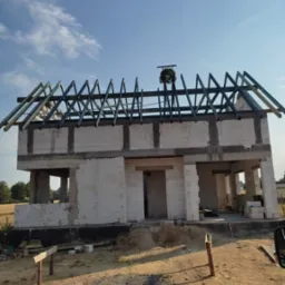 Surowy stan domu z betonu komórkowego z częściowo wykonaną więźbą dachową, widoczny wieniec dożynkowy na szczycie, na pierwszym planie piaszczysty teren budowy.