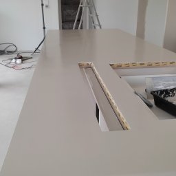 KDE Construction - Jasna, gładka powierzchnia blatu z wyciętymi otworami na oświetlenie LED, zabezpieczonymi taśmą malarską. W tle drabina i sprzęt do nagrywania w jasnym wnętrzu.