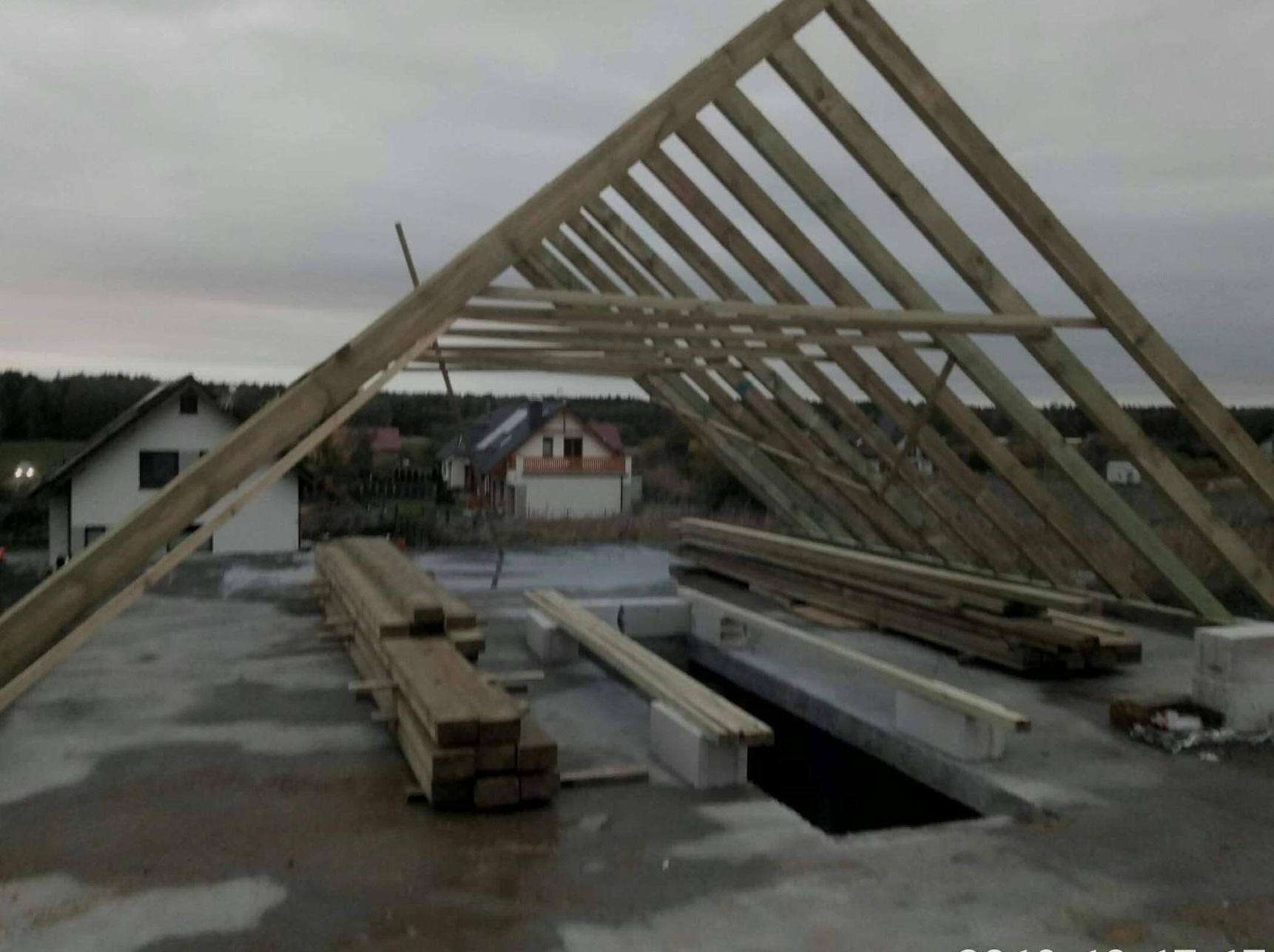 Konstrukcja drewniana dachu w trakcie montażu, widoczne elementy więźby dachowej, z tyłu domy w zabudowie jednorodzinnej, pochmurne niebo.