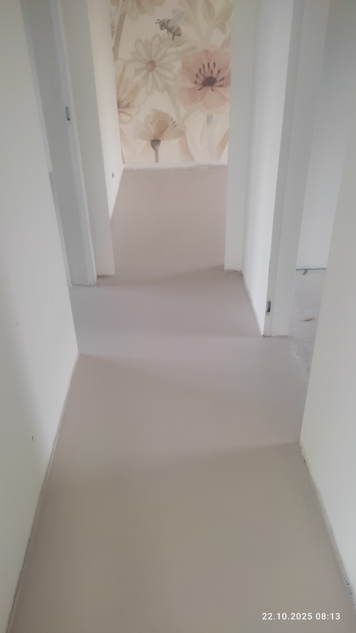 Jasna, gładka posadzka w korytarzu z białymi ścianami i tapetą w kwiaty w tle. Widok z góry, neutralne oświetlenie, świeżo wylana wylewka.