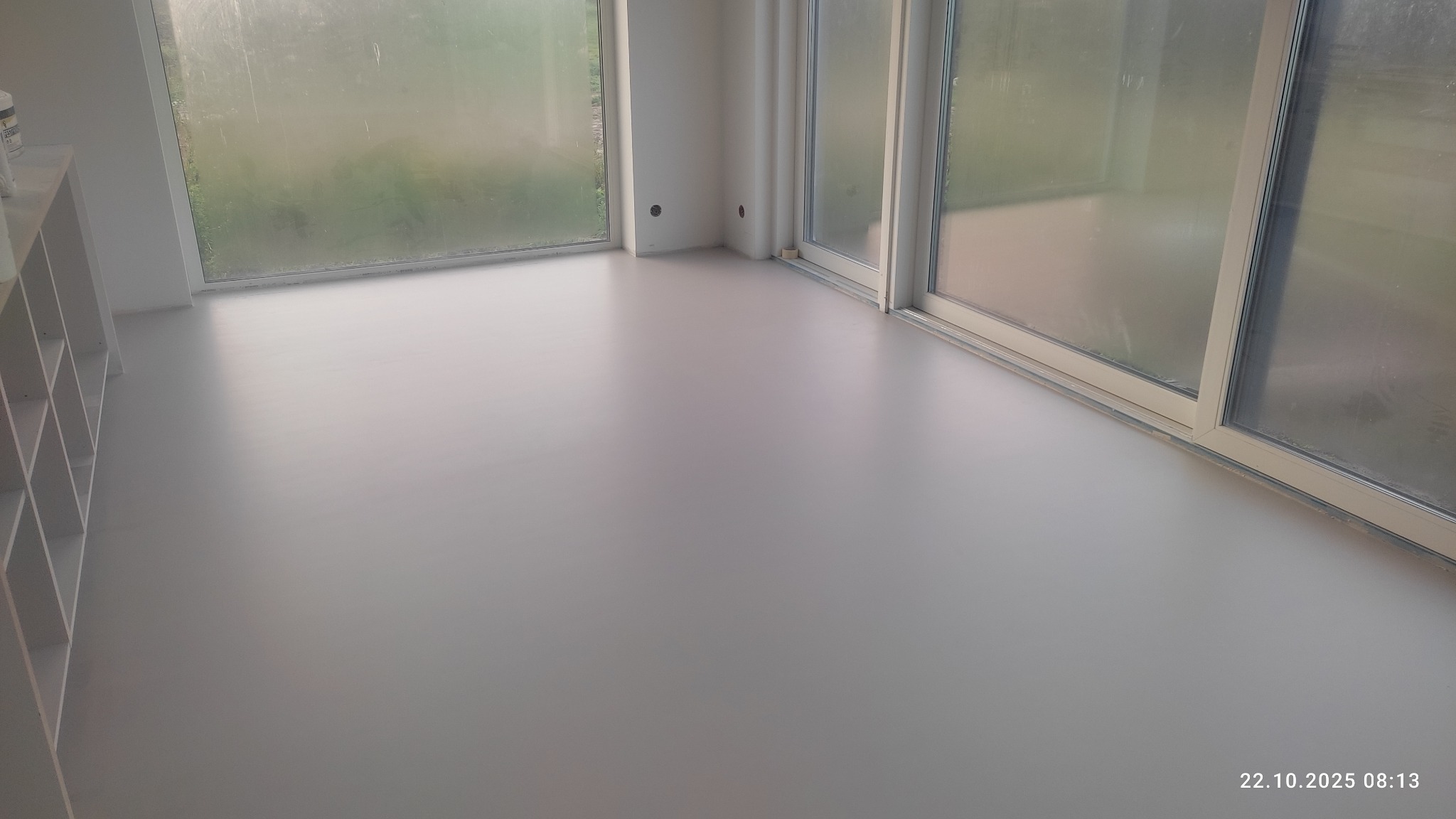 Jasnoszara, gładka posadzka w pokoju z dużymi oknami. Widoczne ślady po wałku malarskim. Minimalistyczny styl, nowoczesne wykończenie.