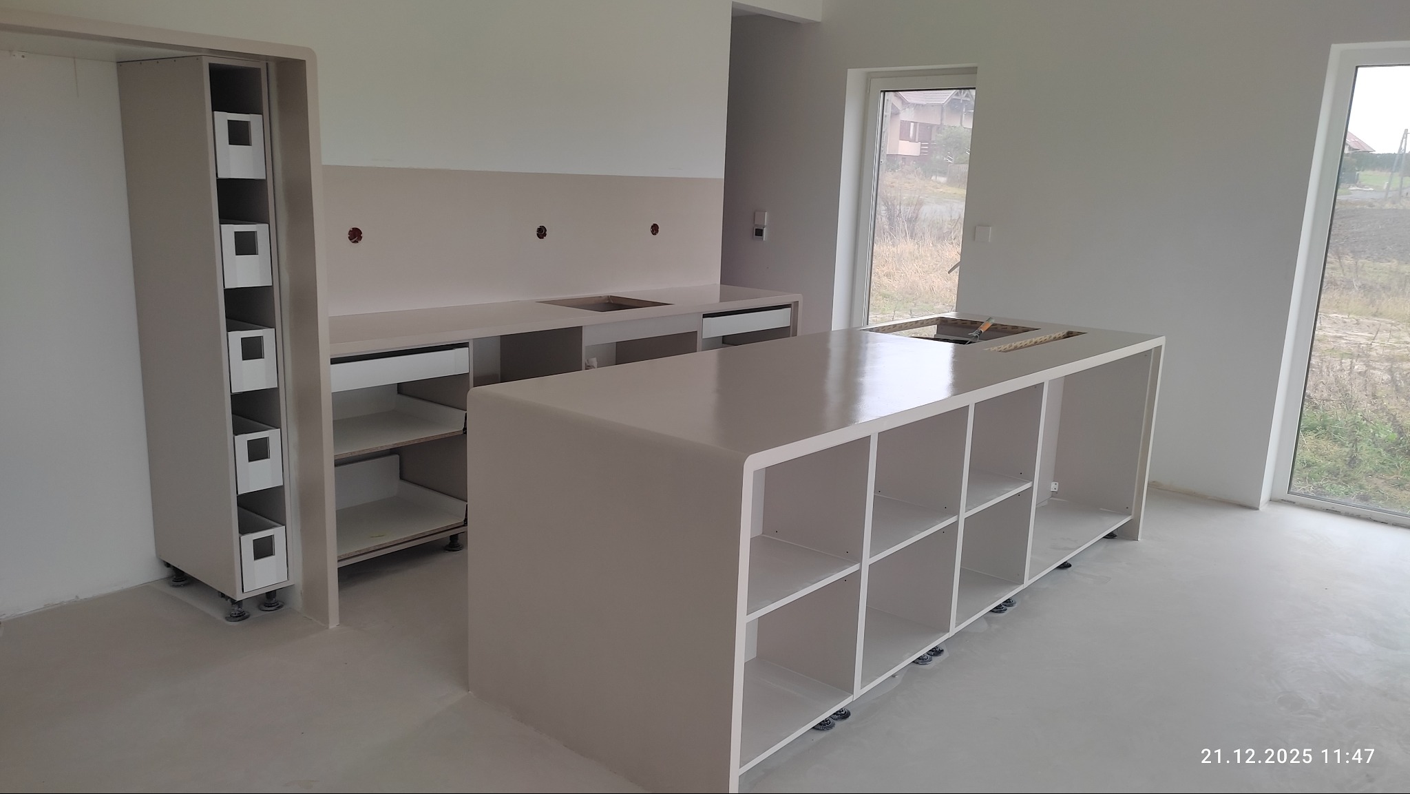 Jasna, minimalistyczna kuchnia w trakcie wykańczania, z wyspą i szafkami bez uchwytów, duże okna, surowy beton na podłodze, widoczne niedoskonałości wykończenia.