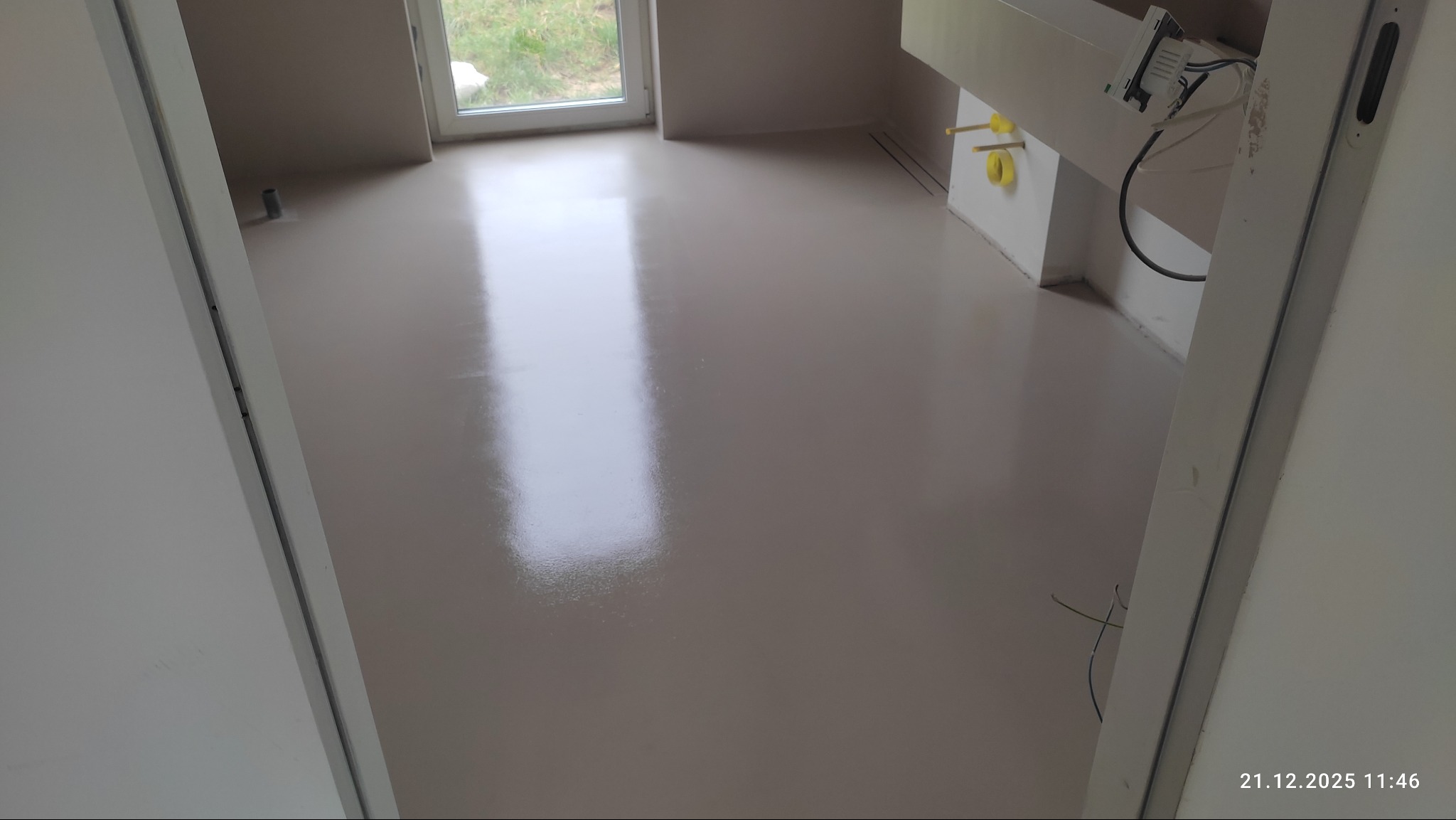 Jasna, gładka posadzka w nowym wnętrzu, widoczna przez otwarte drzwi. Minimalistyczny design, świeżo wylana powierzchnia z delikatnym połyskiem, instalacje w tle.