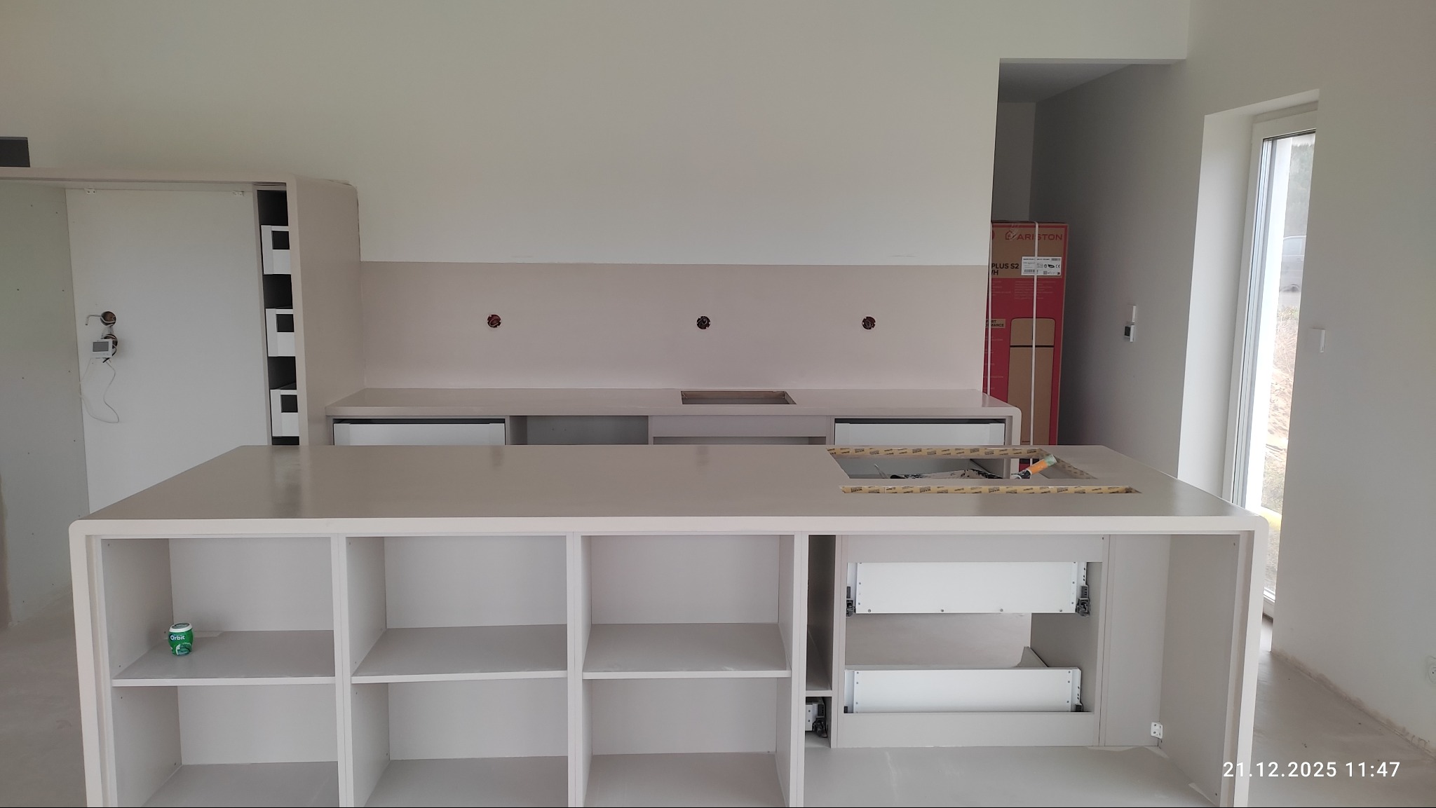 Jasna kuchnia w trakcie montażu: wyspa z otwartymi półkami i szufladami, widoczne otwory na armaturę, minimalistyczny design, surowe wnętrze.