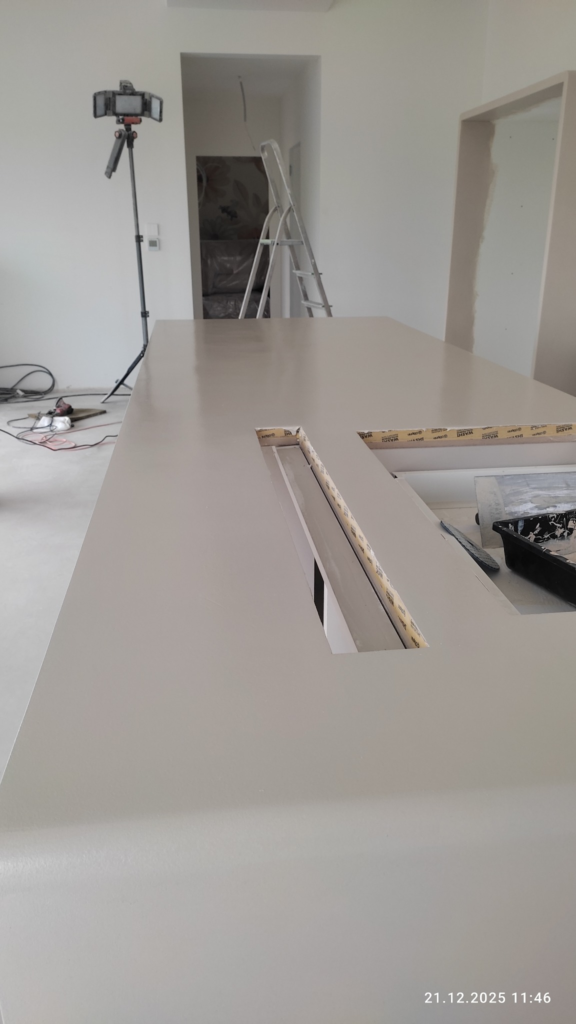 Jasna, gładka powierzchnia blatu z wyciętymi otworami na oświetlenie LED, zabezpieczonymi taśmą malarską. W tle drabina i sprzęt do nagrywania w jasnym wnętrzu.