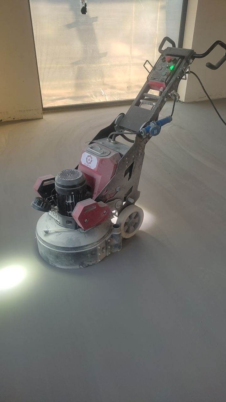 Szlifierka do posadzek w trakcie pracy na gładkiej, szarej powierzchni. Widoczne światło robocze i elementy sterujące maszyny. W tle okno z folią ochronną.