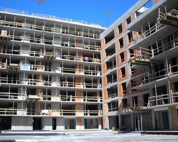 Wielopiętrowy budynek w trakcie budowy z widocznymi szalunkami i rusztowaniami, ceglanymi ścianami działowymi oraz betonowymi elementami konstrukcyjnymi, sfotografowany w słoneczny dzień.