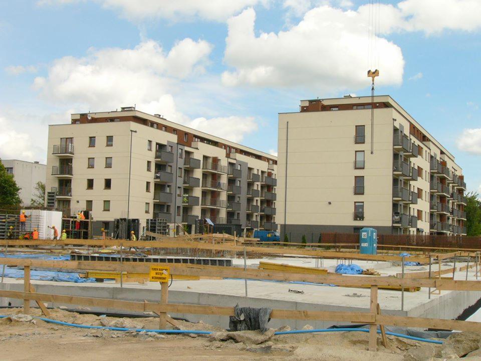 Wczesny etap budowy wielorodzinnego budynku mieszkalnego z balkonami; widoczny plac budowy z fundamentami i pracownikami w kamizelkach odblaskowych, dźwig w tle.
