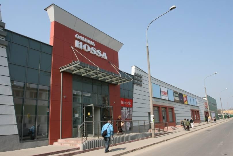 Fasada budynku usługowego Galeria Hossa z czerwonymi i szarymi panelami, przeszklonym wejściem i metalowym daszkiem, widoczna z poziomu ulicy z przechodzącymi osobami i latarniami.