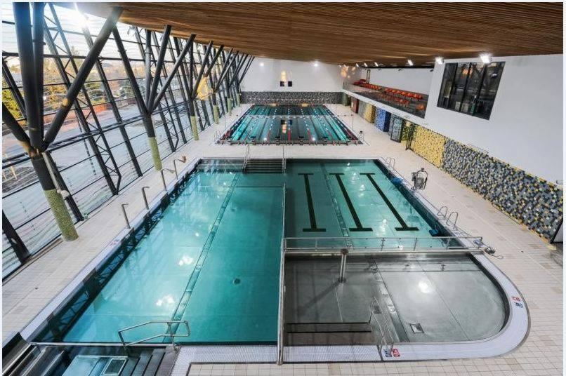 Wnętrze nowoczesnej hali basenowej z dużym przeszkleniem, widok z góry na baseny sportowe z czarnymi pasami na dnie, drewniany sufit i trybuny dla widzów.
