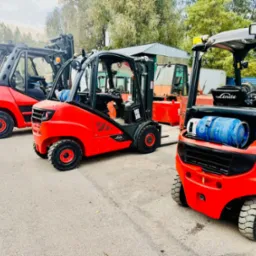 Rząd czerwonych wózków widłowych Linde, z niebieskimi butlami LPG na przednim wózku, stojących na betonowym placu z drzewami w tle.