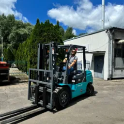 Mężczyzna w okularach przeciwsłonecznych siedzi za sterami turkusowego wózka widłowego marki TCF na tle białego budynku z kominem i błękitnego nieba z chmurami.