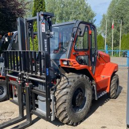 P.U.P.H. TECHTRANS Sp. z o. o. - Pomarańczowy wózek widłowy terenowy z czarnym masztem i widłami, widoczne opony terenowe, na zewnątrz, w tle drzewa i flaga.