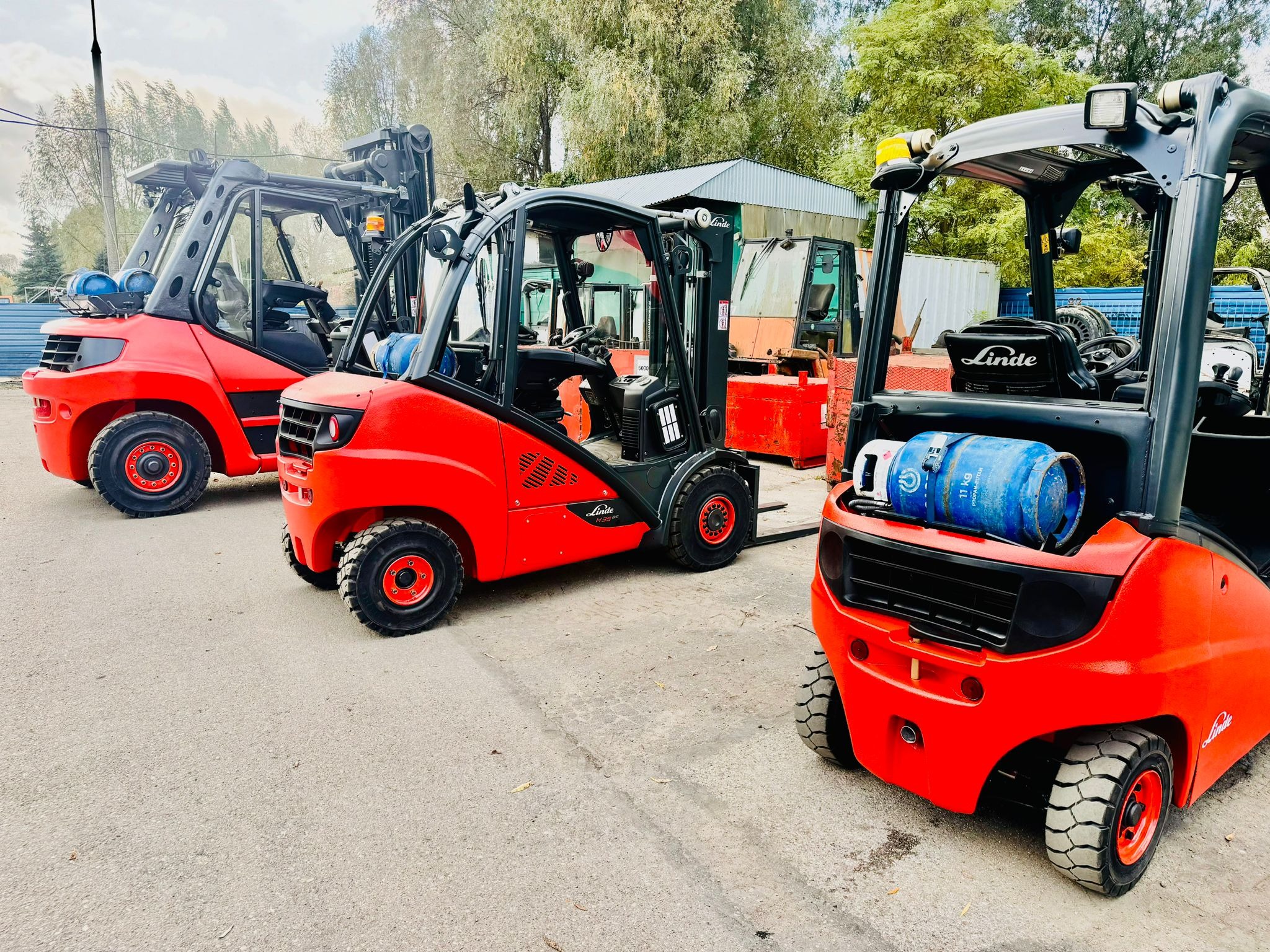Rząd czerwonych wózków widłowych Linde, z niebieskimi butlami LPG na przednim wózku, stojących na betonowym placu z drzewami w tle.