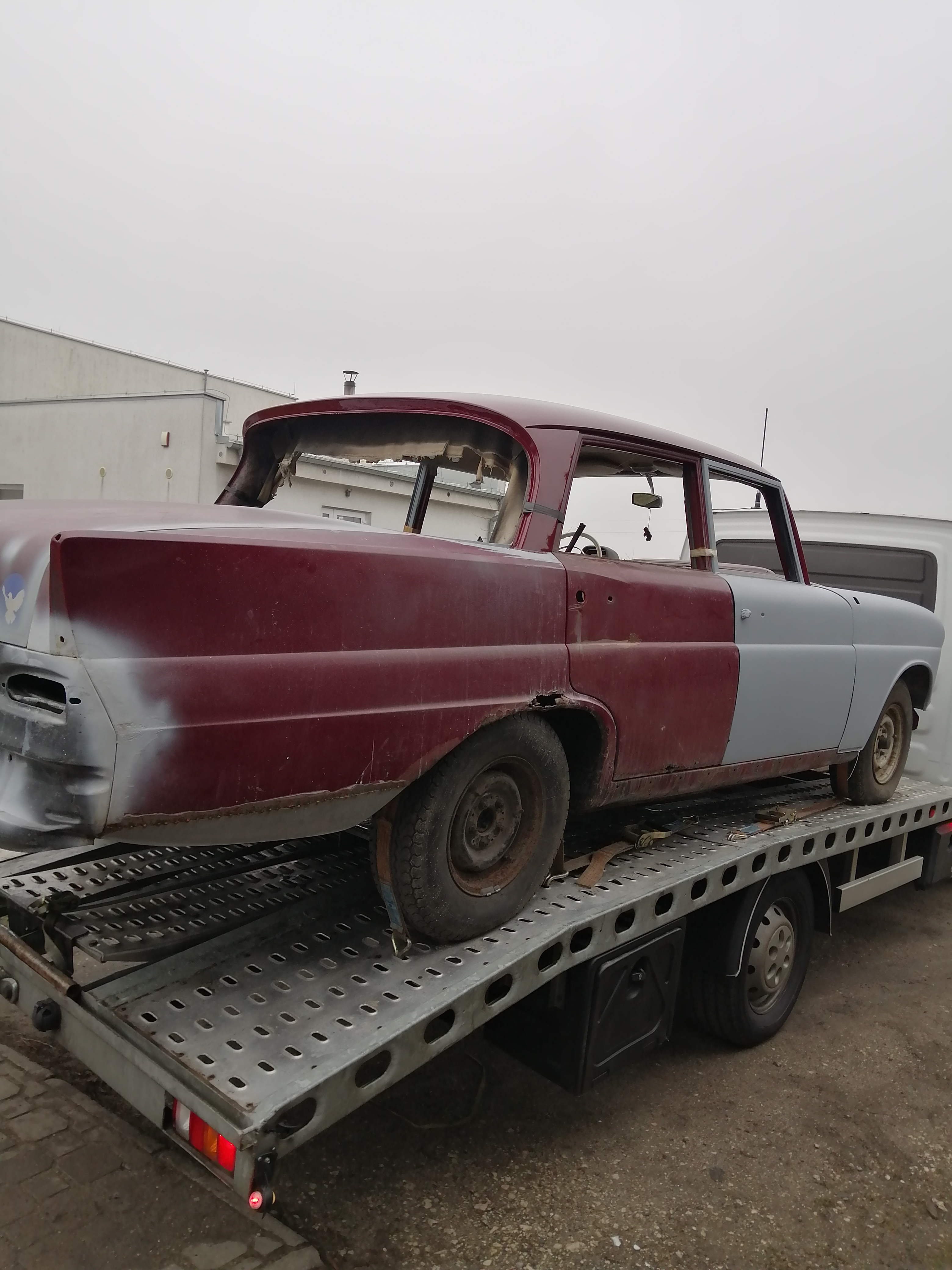 Częściowo odrestaurowane nadwozie klasycznego samochodu Mercedes, w kolorze bordowo-szarym, umieszczone na platformie lawety, widoczne ślady korozji i napraw blacharskich.