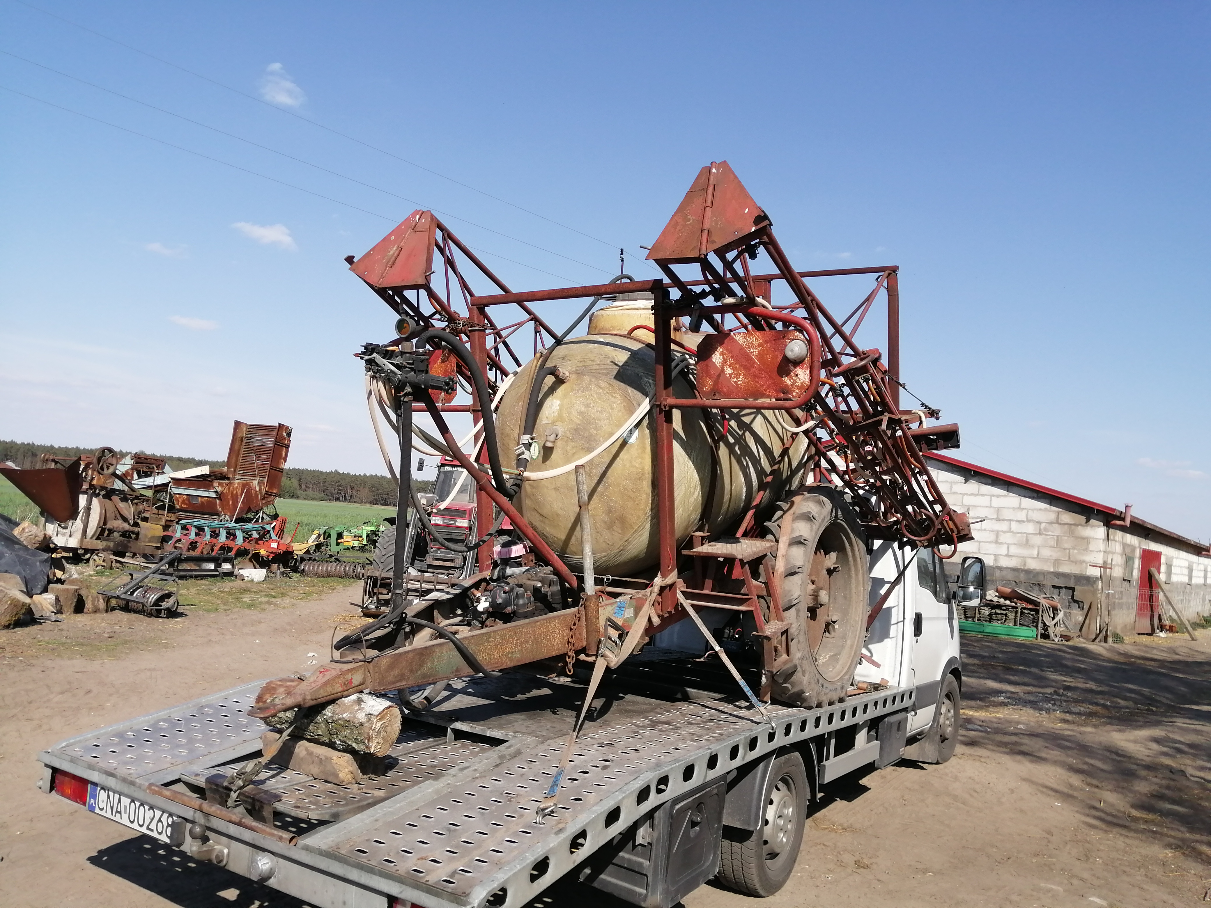 Laweta przewozi stary, rdzewiejący opryskiwacz rolniczy z żółtym zbiornikiem i czerwoną ramą; na platformie leżą drewniane klocki zabezpieczające ładunek.