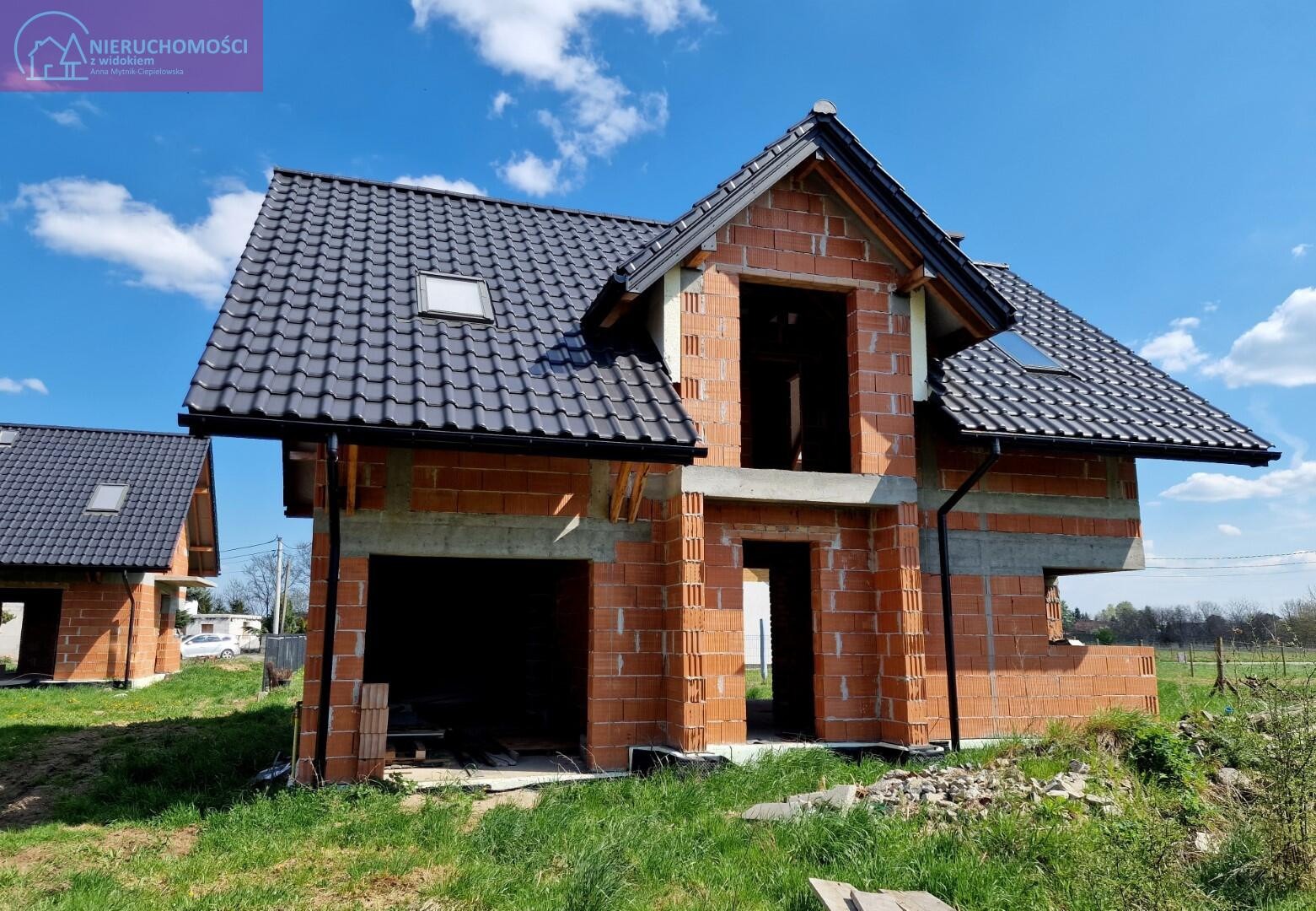 Surowy budynek jednorodzinny w trakcie budowy, z widoczną konstrukcją z czerwonej cegły, czarnym dachem z lukarnami i nieotynkowanymi ścianami, otoczony zieloną trawą i błękitnym niebem z chmurami.