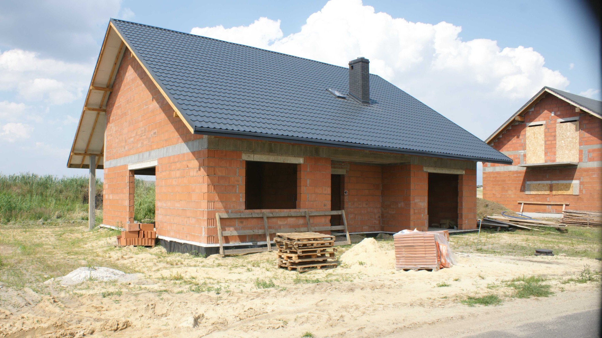 Surowy stan budynku jednorodzinnego z czerwonej cegły z ciemną dachówką, widoczne palety i cegły na piaszczystym terenie, w tle drugi budynek z zabitymi deskami oknami.