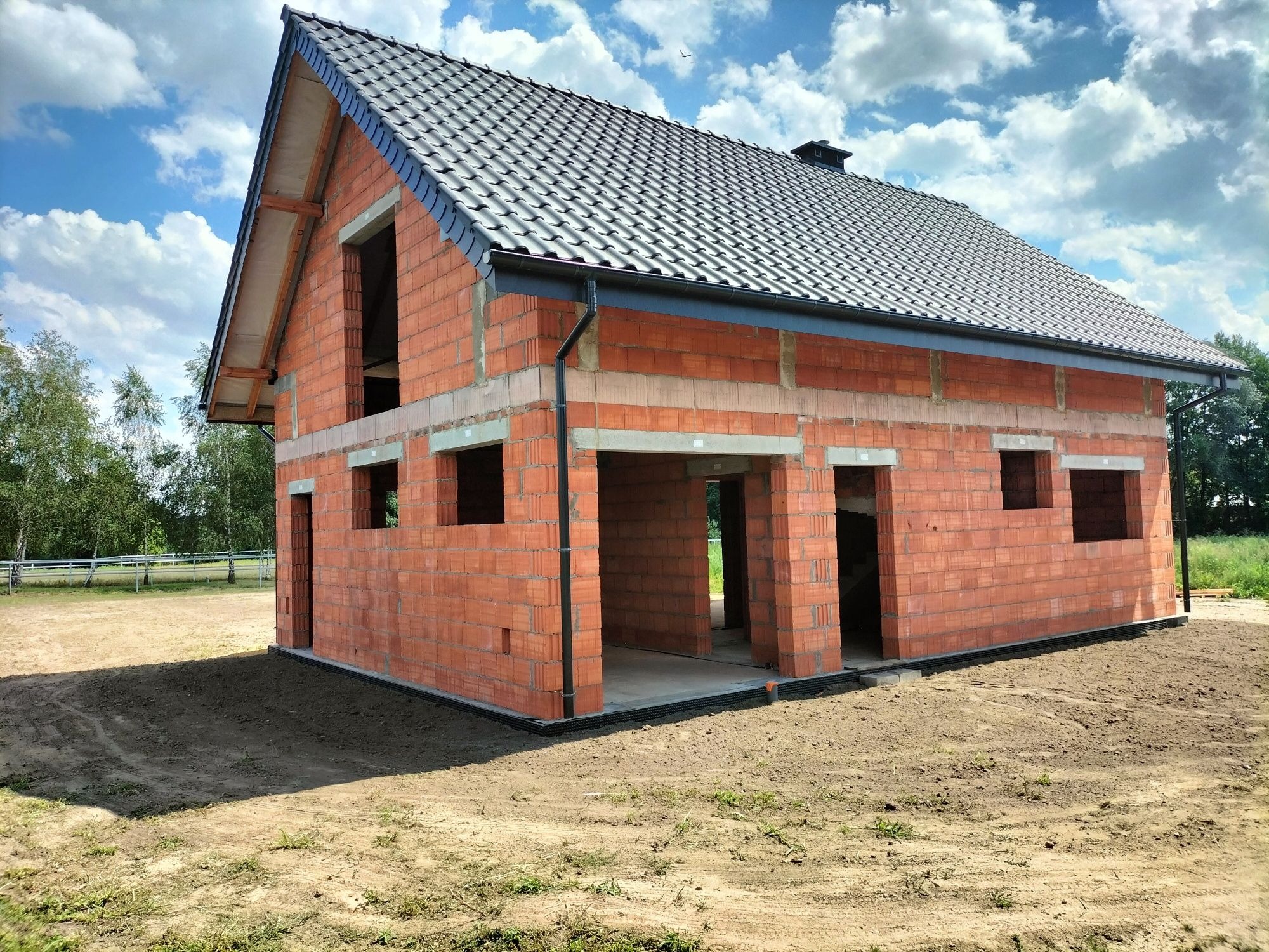 Surowy stan budynku jednorodzinnego z czerwonej cegły, z widoczną czarną dachówką i nieotynkowanymi ścianami, na tle błękitnego nieba z chmurami.