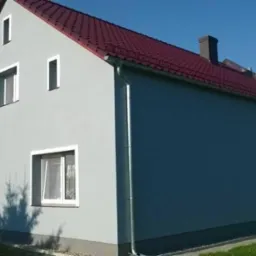 Dwukondygnacyjny dom z szarą elewacją, czerwoną dachówką i białymi oknami, widok z boku na tle błękitnego nieba.