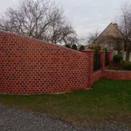 Ceglany, łukowaty mur z ozdobnymi słupkami i metalowym ogrodzeniem, otaczający rabatę z krzewami, na tle domu i pochmurnego nieba.