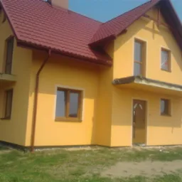 Żółty dom jednorodzinny z czerwoną dachówką, widok z boku, z widocznymi oknami i drzwiami. Częściowo wykończona elewacja z widocznymi elementami zabezpieczającymi.