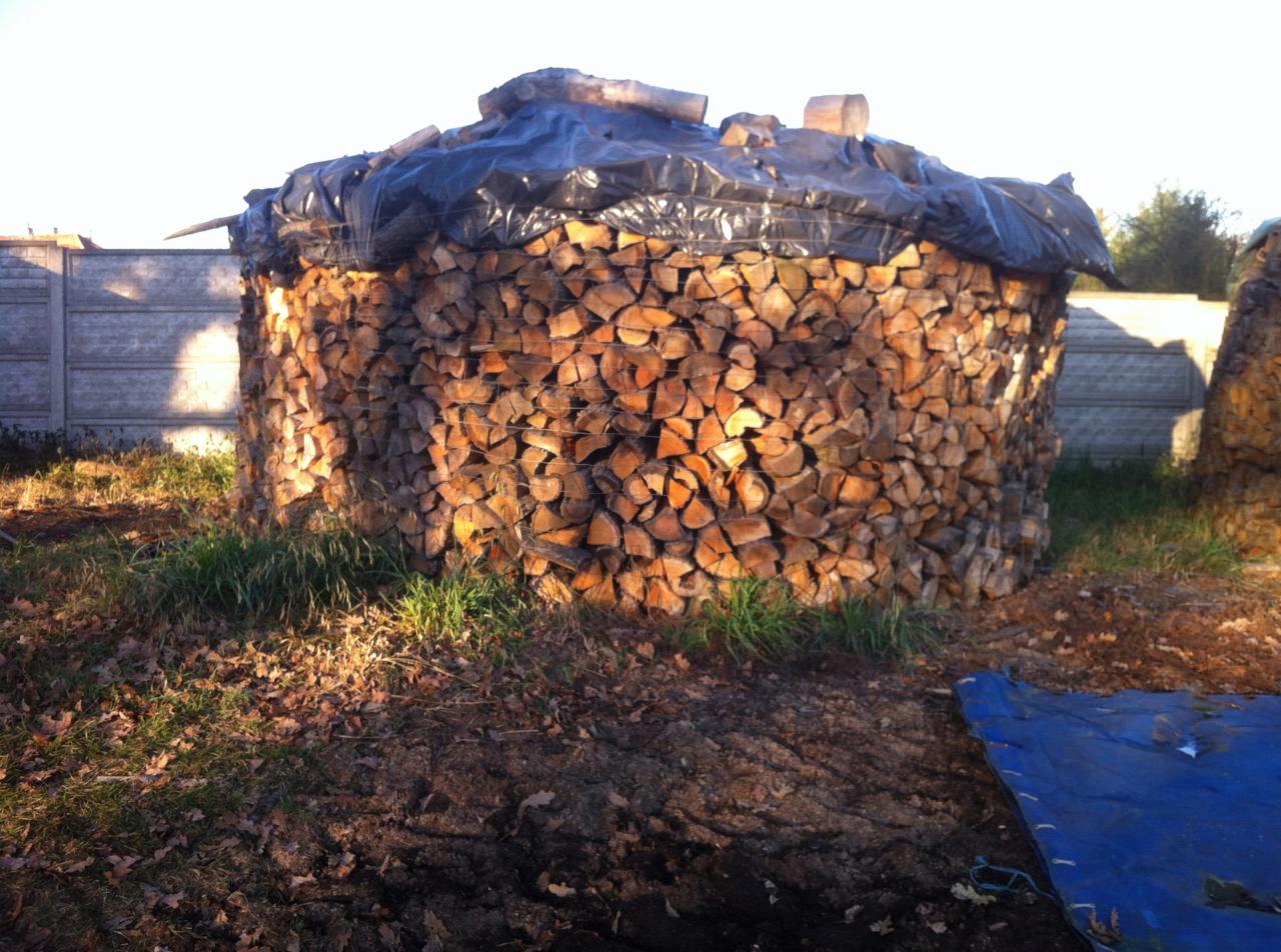 Okrągły stos drewna kominkowego, zabezpieczony czarną folią na wierzchu, obok betonowe ogrodzenie i trawa. Ujęcie w plenerze, w słoneczny dzień.