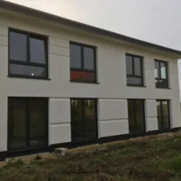 Nowoczesny, dwukondygnacyjny dom z białymi ścianami i ciemnymi ramami okiennymi, z widocznymi panelami dekoracyjnymi na elewacji, w trakcie prac wykończeniowych na zewnątrz, z widocznym fragmentem...