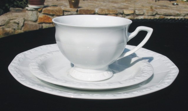 Biała filiżanka z podstawką i talerzem deserowym, porcelana z delikatnym tłoczeniem na krawędziach, ustawiona na czarnym blacie na tle kamiennego muru.