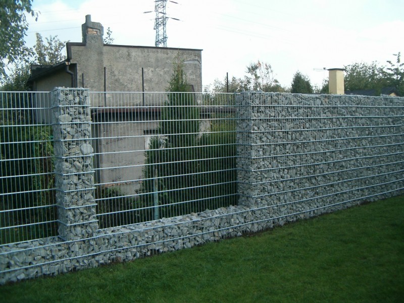 Ogrodzenie gabionowe z szarego kamienia w metalowej siatce, widok z zewnątrz na tle zielonego trawnika i budynku z kominem.