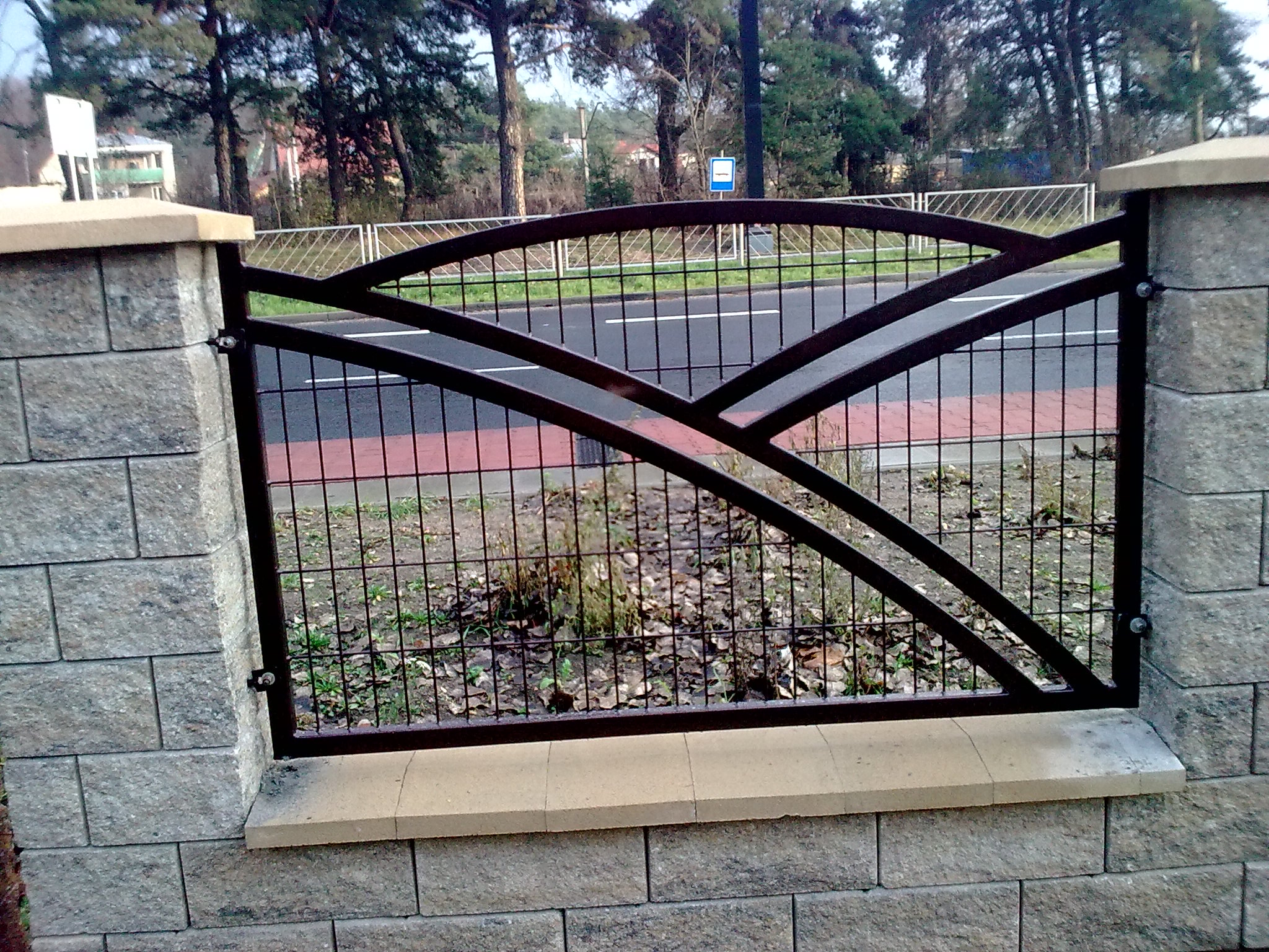 Fragment ogrodzenia z metalową kratą o dekoracyjnych, łukowatych elementach, osadzonego pomiędzy dwoma słupkami z szarej kostki betonowej, widok na ulicę i drzewa w tle.