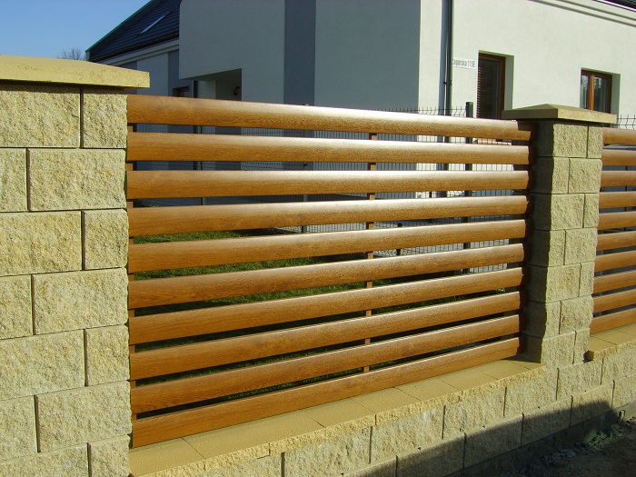 Fragment ogrodzenia z poziomych, brązowych paneli imitujących drewno, osadzonych między słupkami z łupanego kamienia w odcieniach beżu, na tle domu jednorodzinnego.