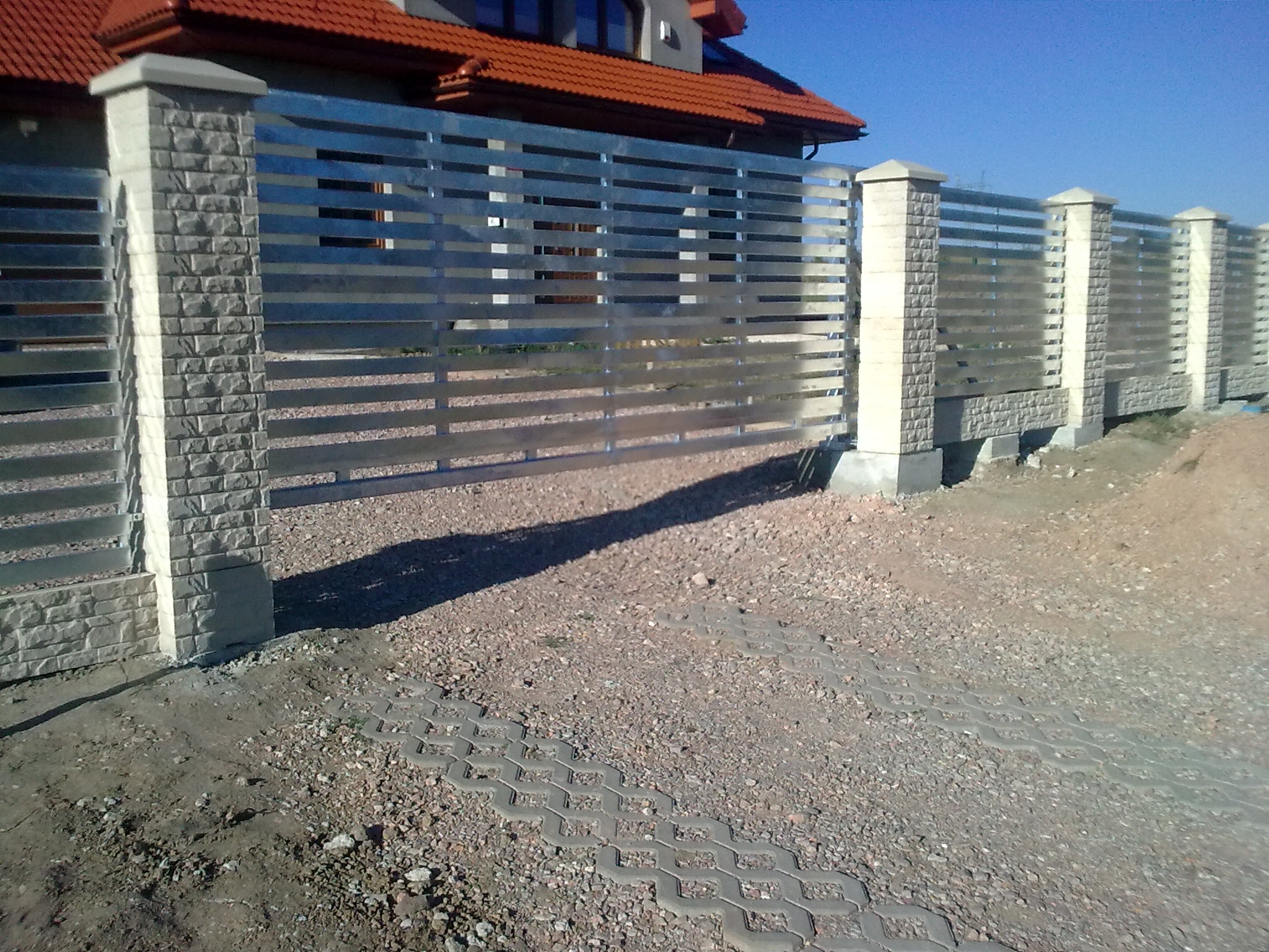 Nowoczesne ogrodzenie poziome z metalowymi przęsłami i kamiennymi słupkami na tle domu z czerwoną dachówką, widok z zewnątrz.