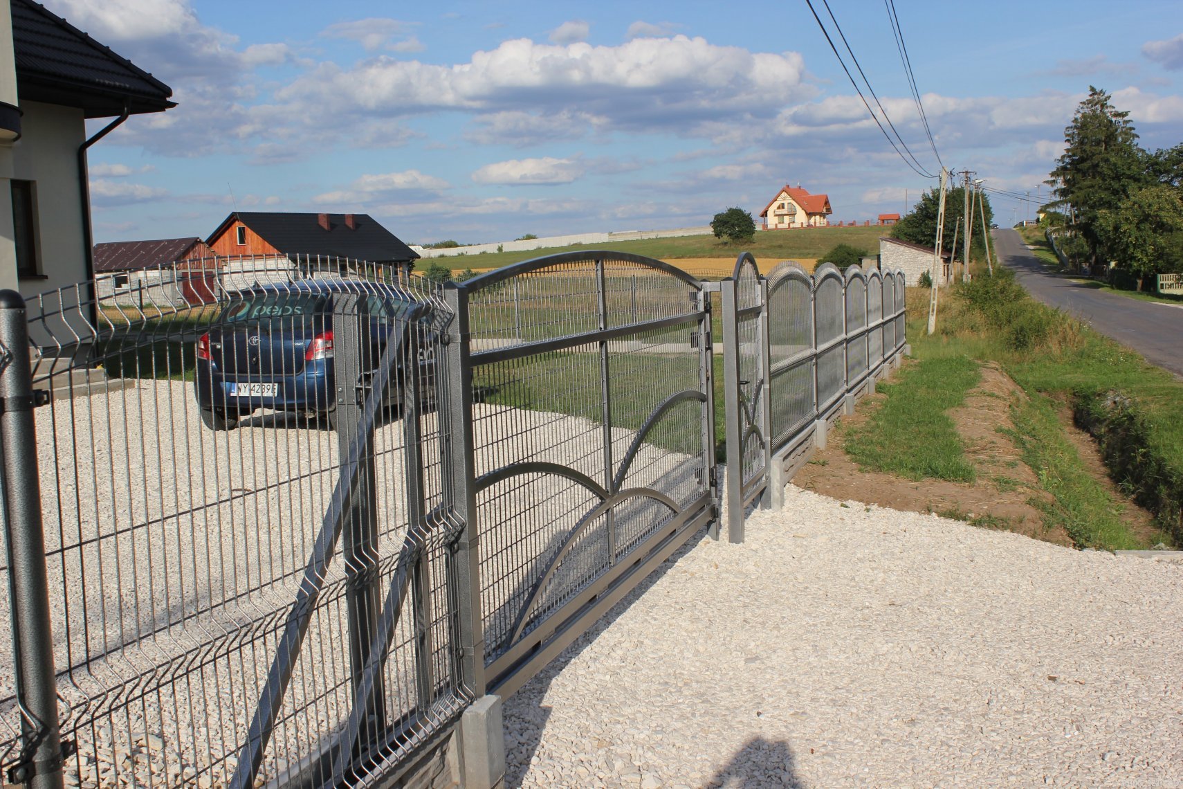 Otwarta, przesuwna brama panelowa z metalową siatką, widok na podjazd z białego żwiru, zaparkowany samochód i domy w tle, słoneczny dzień.