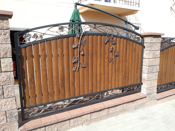 Metalowa brama z drewnianym wypełnieniem i ozdobnymi elementami roślinnymi, zamontowana na kamiennych słupkach, widok z zewnątrz.