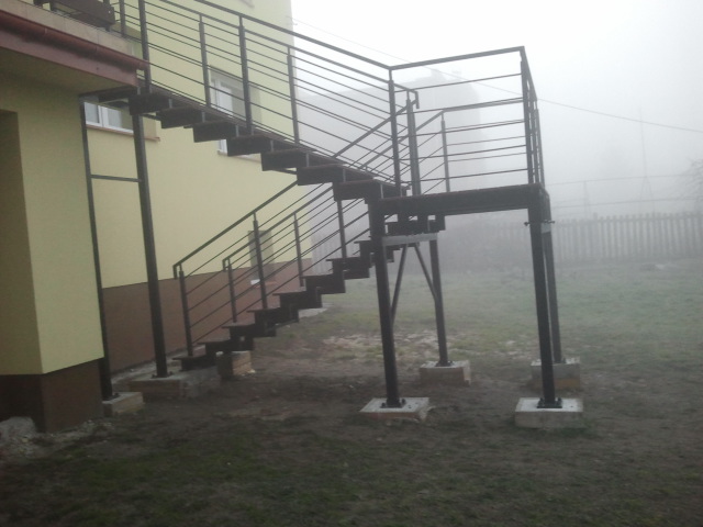 Zewnętrzne schody metalowe z balustradą, prowadzące do budynku, widoczne na tle mglistego krajobrazu.