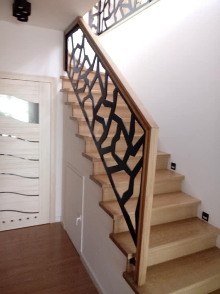 Schody drewniane z ozdobną, czarną, metalową balustradą o abstrakcyjnym wzorze w domu w Kielcach. Widoczna drewniana podłoga i białe drzwi z delikatnym zdobieniem.
