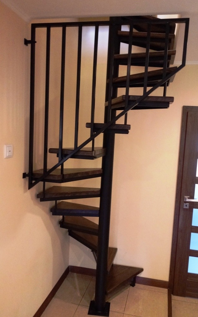 Kompaktowe, spiralne schody z drewnianymi stopniami i czarną metalową konstrukcją, zamontowane w rogu pomieszczenia z beżowymi ścianami i brązowymi drzwiami.