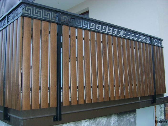 Barierka balkonowa z pionowych desek w odcieniach brązu, zwieńczona czarną metalową ramą z motywem meandru greckiego.