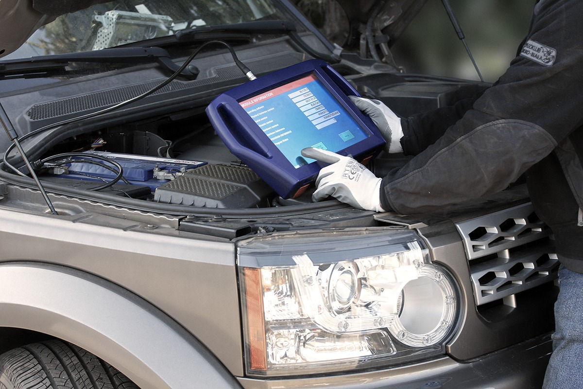 Diagnostyka komputerowa samochodu: osoba w rękawicach roboczych używa tabletu diagnostycznego podłączonego do otwartej maski srebrnego SUV-a, widoczny akumulator i elementy silnika.