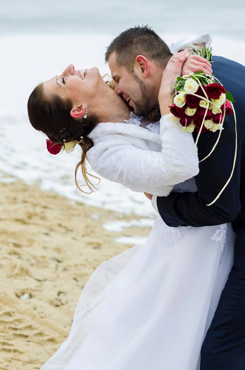 Rozesmiana panna młoda w białej sukni i futerku, obejmowana przez pana młodego w granatowym garniturze na plaży, z bukietem czerwono-białych róż.