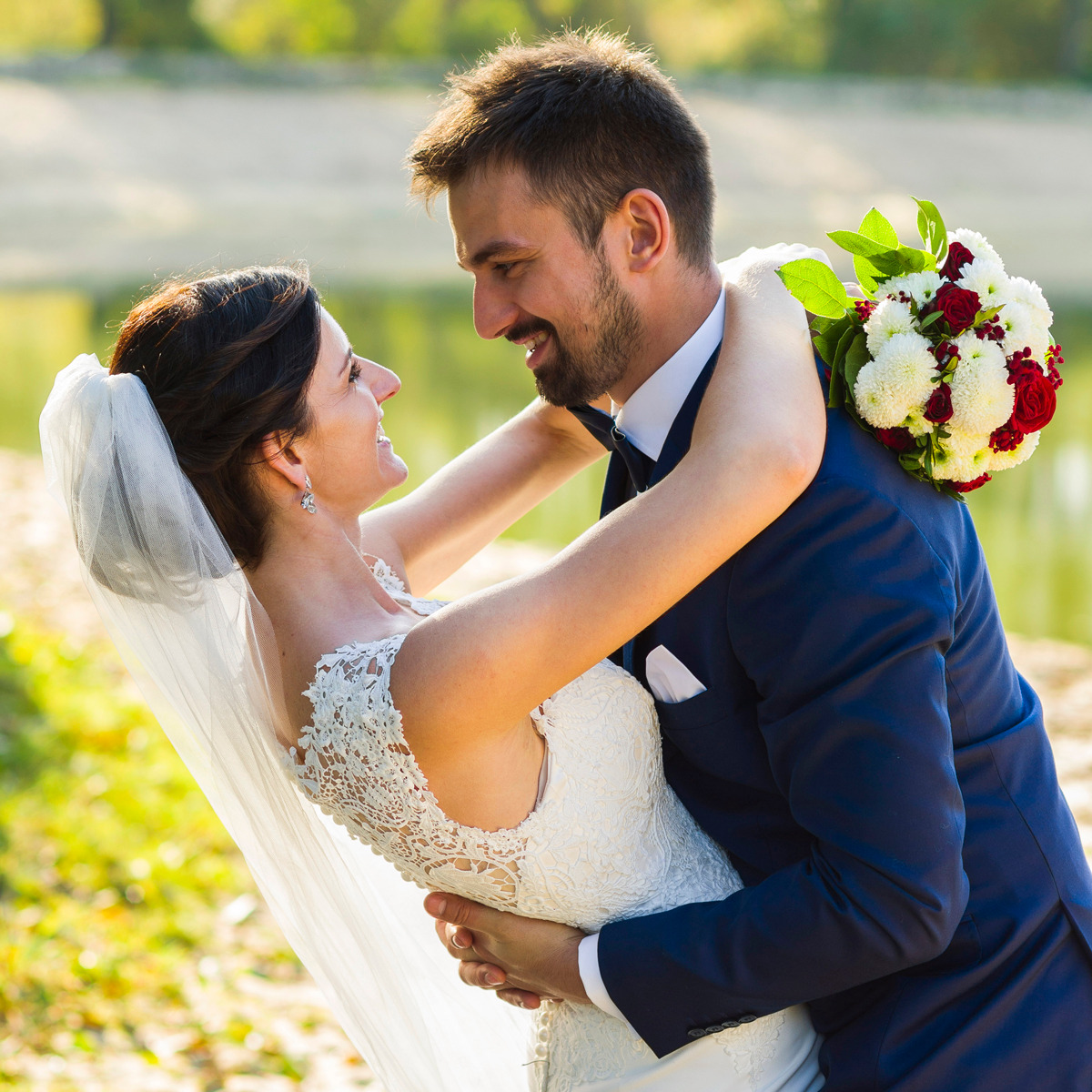 Uśmiechnięta para młoda w objęciach na tle jeziora, panna młoda w koronkowej sukni z welonem, pan młody w granatowym garniturze, bukiet czerwono-białych kwiatów.