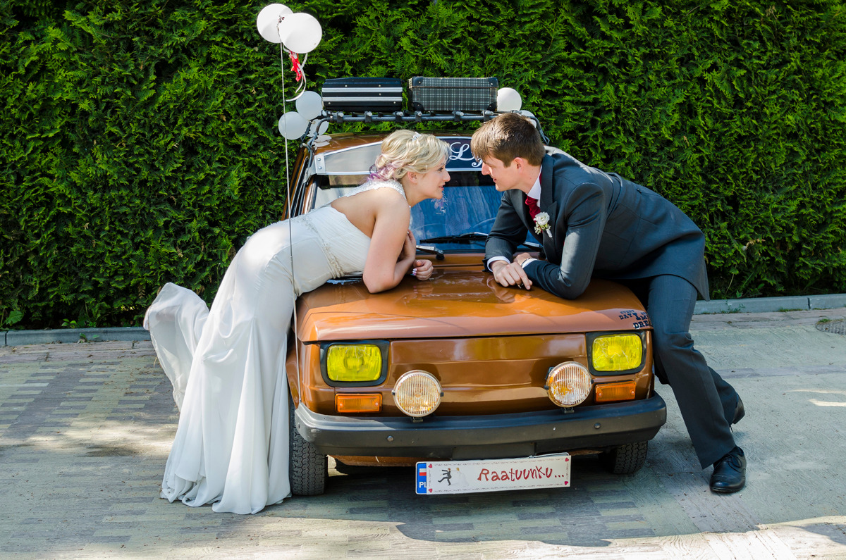 Para młoda w strojach ślubnych opiera się o brązowy samochód z bagażnikiem na dachu i balonami, na tle zielonego żywopłotu.