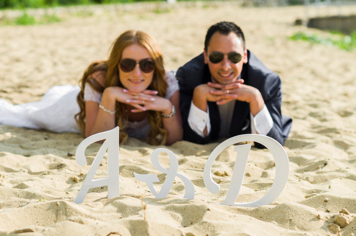 Uśmiechnięta para młoda w okularach przeciwsłonecznych leży na piaszczystej plaży za białymi literami A&D.