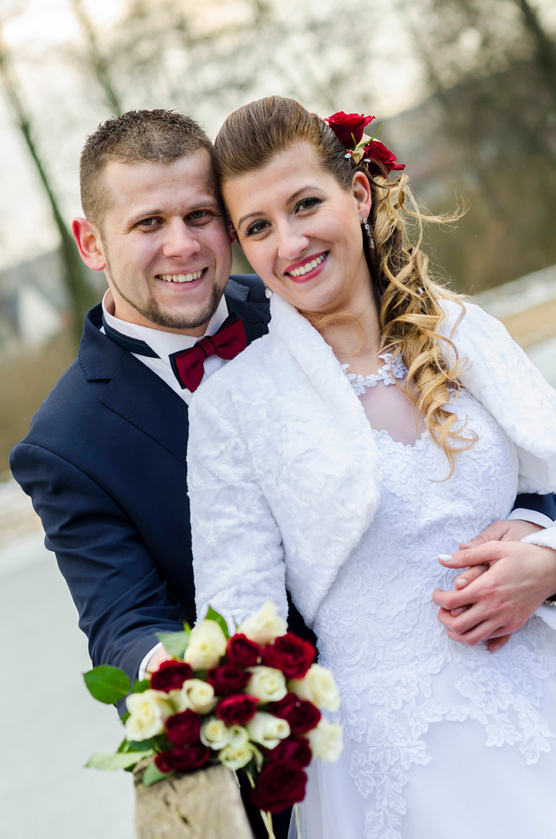 Uśmiechnięta para młoda w strojach ślubnych, panna młoda z czerwonymi różami we włosach i bukietem czerwono-białych róż, pan młody w granatowym garniturze z bordową muchą.