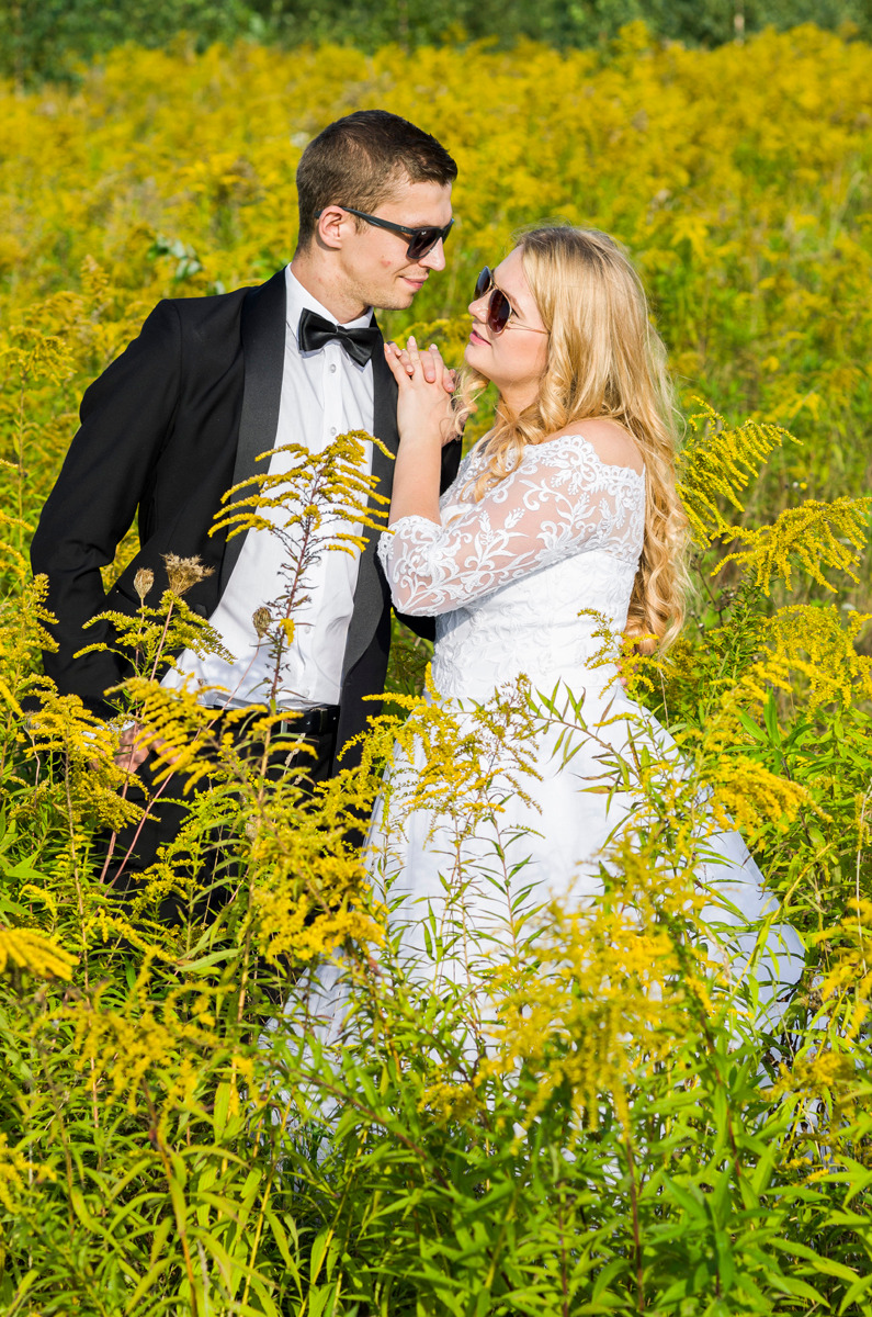 Para młoda w okularach przeciwsłonecznych, stojąca w polu żółtych kwiatów. Panna młoda w koronkowej sukni, pan młody w smokingu z muchą.