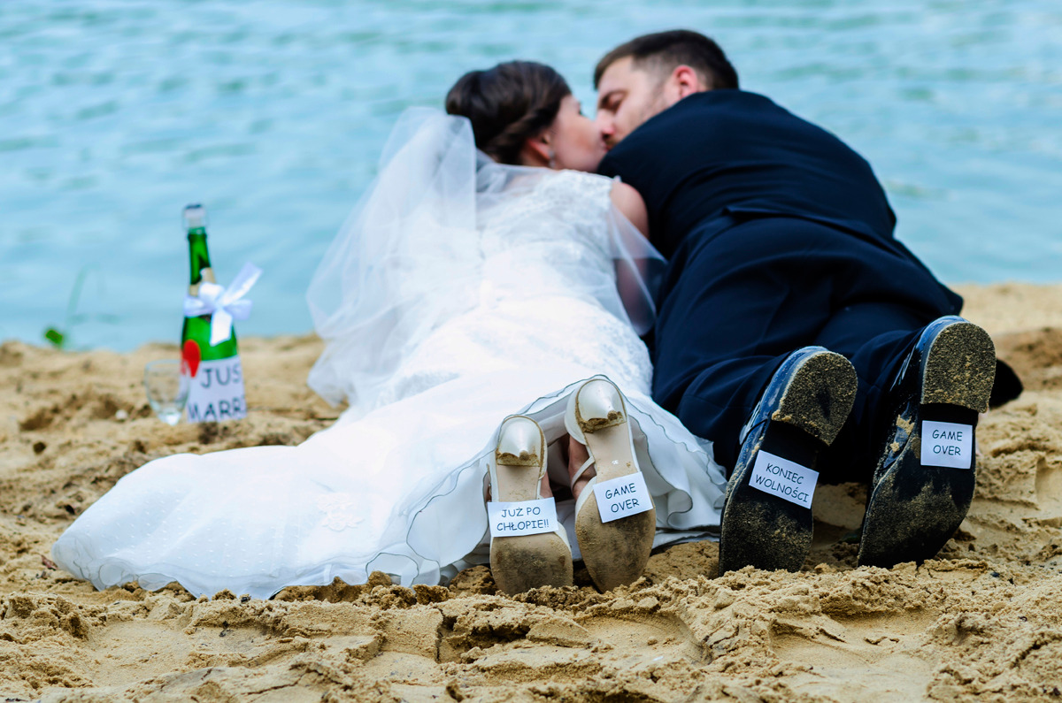 Plażowe ujęcie pary młodej leżącej na piasku, z butami z przymocowanymi kartkami z napisem 'Game Over', 'Już po chłopie' i 'Koniec wolności', butelka szampana z napisem 'Just Married' w tle.